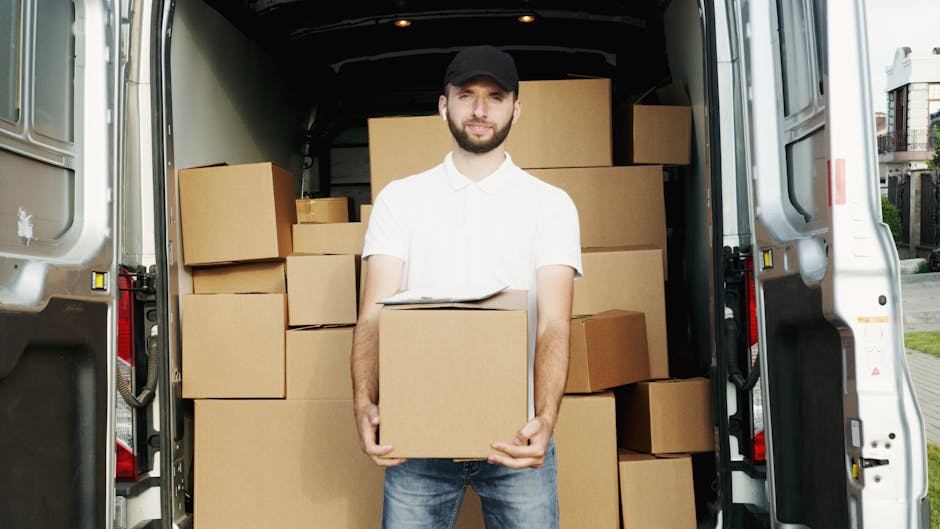 A man wearing a white shirt and black cap stands inside the open rear of a white van, which is loaded with numerous cardboard moving boxes of various sizes. The boxes are packed closely together, some stacked on top of each other, and are wrapped in clear plastic and packing materials. The man is holding a medium-sized box at waist height, with a clipboard resting on top of it, likely for inventory or moving checklists. The scene is set outdoors on pavement near residential buildings, with natural daylight illuminating the area, indicating a home relocation or furniture transport scenario. This image illustrates the loading process involved in professional removals, with Man with Van Kentish Town providing reliable moving services for house removals around Kentish Town, ensuring careful packing and transportation of household items.