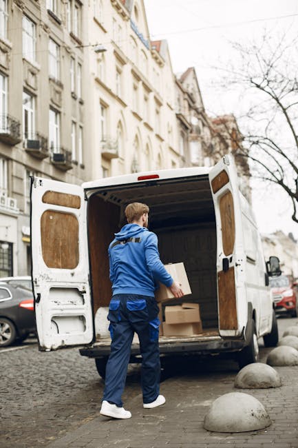 Two movers from Man with Van Kentish Town are engaged in a home relocation process, standing at the rear of a white panel van with its side doors open on a paved street. They are lifting a large, dark green upholstered piece of furniture, likely a sofa or armchair, into the van. The interior of the van is visible, showing a spacious cargo area designed for furniture transport, with some cardboard boxes and packing materials nearby. The weather appears clear and sunny, with a street sign, electrical pole, and part of a sidewalk visible in the background, indicating an urban environment. The movers are dressed in casual workwear, including dark clothing and sneakers, and are focused on the loading process, which involves careful handling and coordination to ensure secure transportation. This scene illustrates professional packing and furniture transport services as part of a comprehensive moving or house removal service offered by Man with Van Kentish Town.