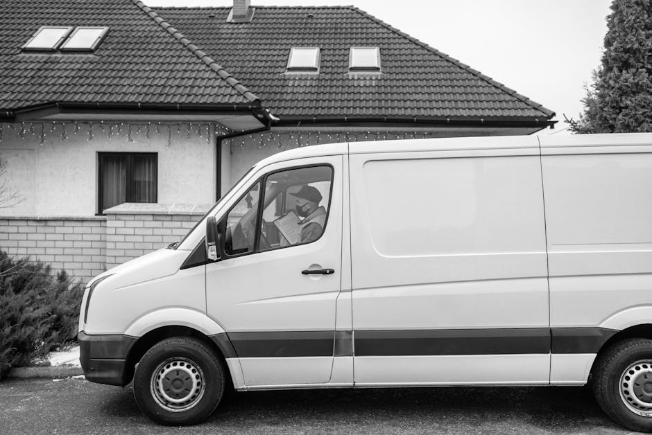 A white cargo van parked on a residential street in front of a house with a tiled roof, small attic windows, and string lights hanging under the eaves. Inside the van, a person wearing a face mask is sitting in the driver's seat, holding a clipboard and looking out through the window. The van is positioned close to the entrance of a driveway or loading area, suggesting it is in the process of a home relocation or furniture transport. The setting includes low shrubs and pavement, with the house's exterior walls made of white plaster and exposed brickwork. The scene captures a typical domestic moving scenario, illustrating the logistics involved in packing and loading belongings for a house removal, consistent with services offered by Man with Van Kentish Town for Victorian-style homes along Fortess Road.