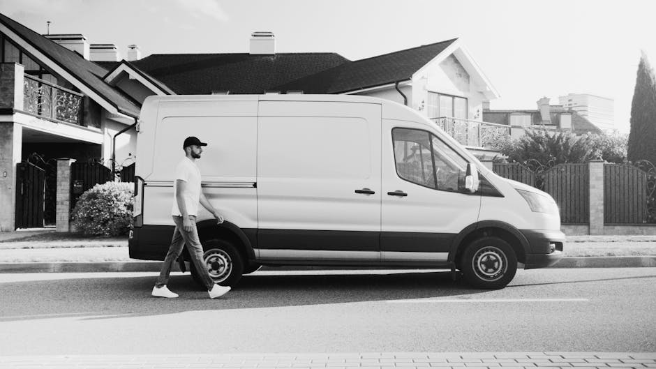 A man wearing a baseball cap, white t-shirt, and jeans is walking past a white van parked on a residential street in front of a row of Victorian houses with decorative railings and balconies. The van, belonging to Man with Van Kentish Town, is positioned parallel to the pavement with the sliding side door closed. The vehicle appears to be used for home relocation or furniture transport, as the setting suggests loading or unloading activities associated with packing and moving. The street is lined with houses featuring pitched roofs, chimneys, and well-maintained gardens, with natural daylight illuminating the scene. The image captures the typical loading process of a house removal, focusing on the van's exterior and the surrounding environment, reflecting professional moving services in a London neighbourhood.