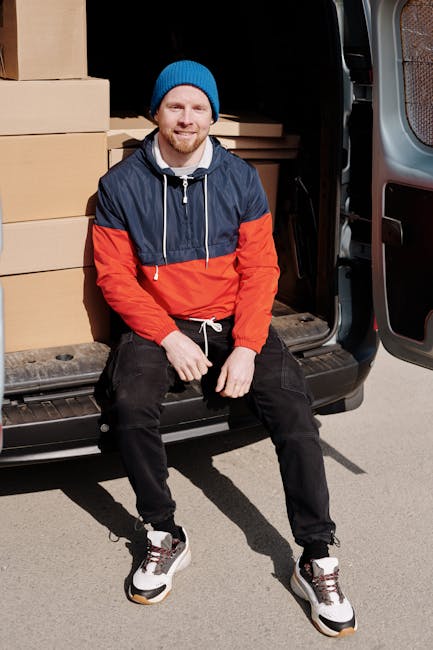 A man with a beard, wearing a blue beanie and a colour-blocked jacket in navy blue, red, and white, is sitting on the edge of an open black van during a home relocation. Inside the van, there are neatly stacked cardboard boxes, some sealed with packing tape, and additional packing materials such as bubble wrap and plastic stretch wrap visible around the boxes. The man is positioned just outside a doorway, with the van parked on a paved surface, suggesting the loading process of furniture transport. The background shows part of a residential building, and natural daylight illuminates the scene. The man appears to be taking a break during packing and loading, representing the logistics involved in professional removals or house moves, as offered by Man with Van Kentish Town. This scene exemplifies the packing, loading, and transportation stages of a property move.