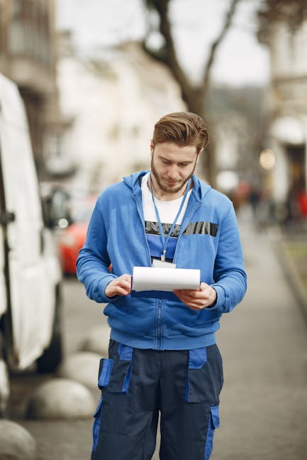 A man dressed in a blue work hoodie and dark blue work trousers, standing outdoors on a paved street near white residential buildings, is reading a clipboard or document. The background includes parked vehicles, trees, and blurred urban scenery. The scene suggests a professional involved in house removals or moving services, possibly reviewing instructions or coordinating a home relocation. Natural daylight illuminates the scene, highlighting the man's focused expression. This image captures a moment of planning and organization in the context of furniture transport or packing and moving processes, with the man representing a member of the team from Man with Van Kentish Town during a house removal or relocation service.
