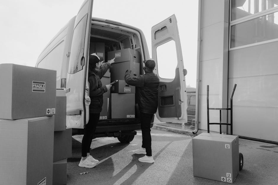 Two movers from Man with Van Kentish Town are engaged in a home relocation process, loading packed cardboard boxes and household furniture into a white cargo van situated outdoors near a modern building. The female mover, wearing casual clothing including a jacket and white sneakers, is inside the van, handing or arranging items on top of larger boxes. The male mover, dressed in a cap, dark jacket, and white sneakers, stands outside facing the open rear doors, assisting with a stack of packed boxes and furniture secured within the vehicle. Several cardboard boxes marked with fragile labels are positioned both inside and outside the van, with some on the pavement and one on a trolley nearby, indicating the careful packing involved in furniture transport and packing and moving procedures. The environment is well-lit with natural daylight, capturing the busy loading process typical of a professional removals service, supporting efficient logistics for a detailed home relocation experience conducted by Man with Van Kentish Town near Kentish Town Station.