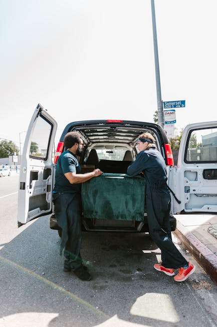 Two movers from Man with Van Kentish Town are loading a large, dark green upholstered armchair into the open rear cargo area of a white van parked on a street. The van's rear doors are wide open, revealing an interior fitted with protective padding and packing materials. One mover, wearing a dark jacket and red sneakers, is holding the armchair steady while the other, dressed in a teal shirt, assists with the lifting. The scene takes place outdoors on a city street with a lamppost, a street sign marked 'Somerset,' and a sidewalk visible in the background. The façade of nearby buildings and a cloudy sky complete the setting, which is part of a home relocation process involving furniture transport and packing for a house move.