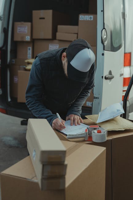 Two movers from Man with Van Kentish Town are engaged in a home relocation process, loading packed cardboard boxes and household furniture into a white cargo van situated outdoors near a modern building. The female mover, wearing casual clothing including a jacket and white sneakers, is inside the van, handing or arranging items on top of larger boxes. The male mover, dressed in a cap, dark jacket, and white sneakers, stands outside facing the open rear doors, assisting with a stack of packed boxes and furniture secured within the vehicle. Several cardboard boxes marked with fragile labels are positioned both inside and outside the van, with some on the pavement and one on a trolley nearby, indicating the careful packing involved in furniture transport and packing and moving procedures. The environment is well-lit with natural daylight, capturing the busy loading process typical of a professional removals service, supporting efficient logistics for a detailed home relocation experience conducted by Man with Van Kentish Town near Kentish Town Station.