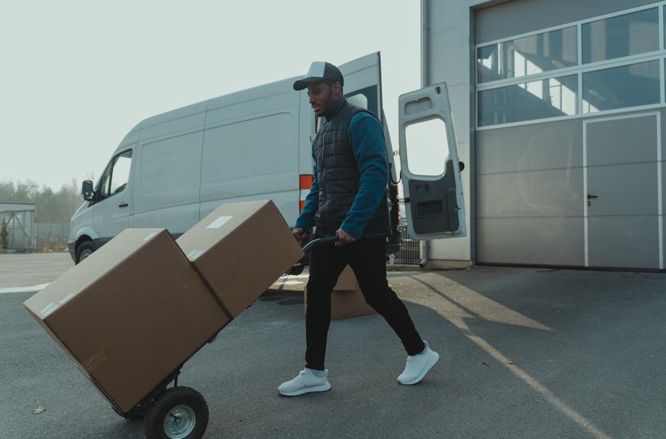 A man wearing a flat cap and dark jacket stands on a street pavement near a small utility vehicle used for home relocation, with cardboard boxes and packing materials on the vehicle's flatbed. Behind 
