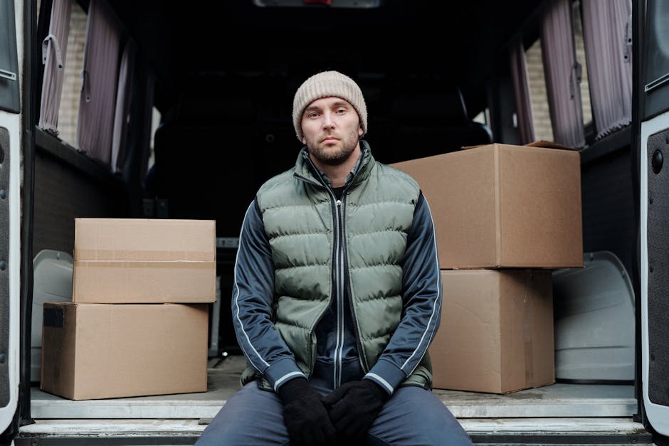 A man with a serious expression is sitting on the pavement just outside the rear of a large moving van, which is loaded with several cardboard boxes of varied sizes. The boxes are packed tightly inside the van, some stacked on top of others, and are made of brown corrugated cardboard, with clear packing tape visible on some. The man is dressed in a beige knitted beanie, a green quilted vest over a navy blue and grey sports jacket, and dark gloves, suggesting chilly weather. The open van door reveals a glimpse of the interior with more packed items, and the van's exterior appears to be grey. The scene captures the loading or unloading process during a home relocation or furniture transport, with the focus on the individual and the packed boxes ready for moving, highlighting the logistics involved in local house removals. This image supports the services offered by Man with Van Kentish Town within their removals category, emphasizing packing and moving procedures associated with small office or shop relocations.