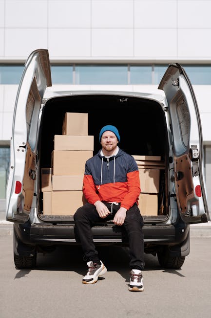 A man wearing a blue beanie, a navy and red jacket, and black pants sits on the edge of an open van's tailgate, with his hands resting on his knees. The van's interior is filled with neatly stacked cardboard boxes of various sizes, covered with plastic wrap for protection during furniture transport and packing for a home relocation. The van is parked on a paved surface in front of a modern building with large glass windows and a tiled exterior, indicating an urban environment typical for house removal services. The background includes a clear sky, and the scene captures the loading process involved in professional moving and furniture transport, with visible equipment such as the van, boxes, and protective packaging materials associated with house removals by Man with Van Kentish Town.