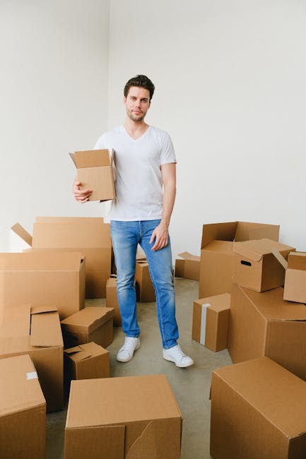 A man in a white t-shirt and blue jeans standing in a room filled with cardboard moving boxes of various sizes, some open and some sealed, arranged on the floor. He is holding a small cardboard box in his right hand and looking towards the camera. The room has white walls and a plain floor, with natural light illuminating the scene. The boxes are organized around him, indicating an ongoing home relocation process, likely as part of furniture transport or packing and moving activities. In the background, a doorway leads to another room, and the scene suggests preparation for a house removal service managed by Man with Van Kentish Town, specializing in house removals and relocation logistics.