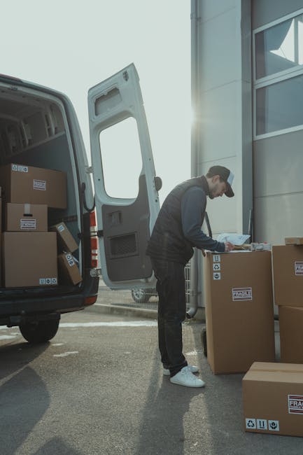 A man wearing a black cap, dark jacket, and white sneakers is standing outdoors near the open back of a white van loaded with cardboard boxes of various sizes. He is holding a clipboard and appears to be recording details or checking items as part of a home relocation process. The boxes are sealed with packing tape and some display labels, indicating they are ready for transport. The van is parked next to a modern building with large glass windows, and several packed boxes are arranged on the pavement nearby. This scene illustrates the loading process involved in furniture transport and packing for house removals, with the man coordinating the logistics of moving belongings from a property. The natural sunlight suggests daytime, and the setting focuses on professional moving services offered by Man with Van Kentish Town, emphasizing careful handling and organized packing during a house relocation.