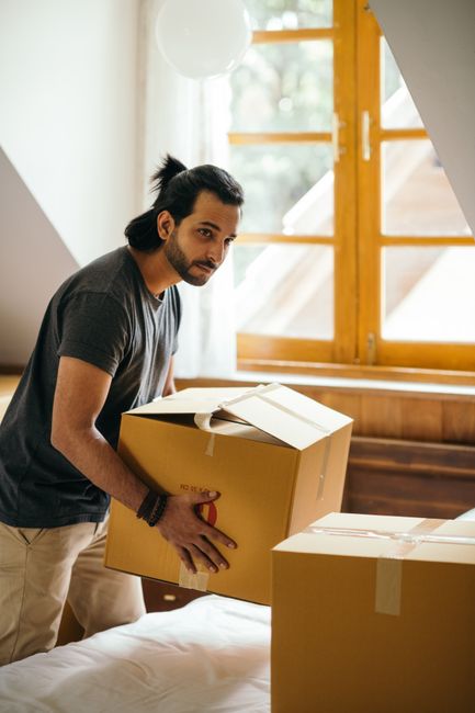 A man in a white t-shirt and blue jeans standing in a room filled with cardboard moving boxes of various sizes, some open and some sealed, arranged on the floor. He is holding a small cardboard box in his right hand and looking towards the camera. The room has white walls and a plain floor, with natural light illuminating the scene. The boxes are organized around him, indicating an ongoing home relocation process, likely as part of furniture transport or packing and moving activities. In the background, a doorway leads to another room, and the scene suggests preparation for a house removal service managed by Man with Van Kentish Town, specializing in house removals and relocation logistics.