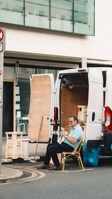 A man with glasses, wearing a light blue t-shirt and dark trousers, is seated on a wooden chair outside a white van parked on a street in Kentish Town. The van's rear doors are open, revealing a partially packed interior with large cardboard boxes, wooden pallets, and packing materials such as plastic wrap and fabric covers. A wooden panel leans against the van's door, and a trolley is positioned nearby. The scene is set in front of a modern building with large glass windows, and a street sign indicating parking restrictions is visible at the top left. The setting captures the process of home relocation, with the man taking a break during the moving operation, supported by visuals of furniture, boxes, and packing supplies involved in furniture transport and packing and moving activities. Man with Van Kentish Town occasionally features for house removal services.