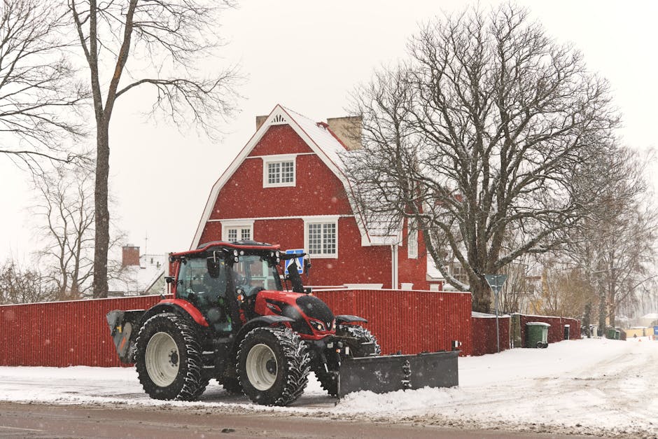 A red tractor equipped with a front-mounted snow plow blade clearing snow from a residential street in front of a red house with white-trimmed windows and a sloped roof. The tractor is parked on the pavement, with its tires partially embedded in the accumulated snow. Behind the tractor, a tall, leafless tree stands next to a wooden fence that runs along the property boundary. Several green and black waste bins are positioned near the fence, and a narrow pathway is visible leading towards the house. The scene is set in winter, with snow falling gently and overcast skies above. This image captures the process of snow removal in a suburban area, supporting the full range of household and moving logistics that Man with Van Kentish Town might coordinate during adverse weather conditions for house relocations and furniture transport.
