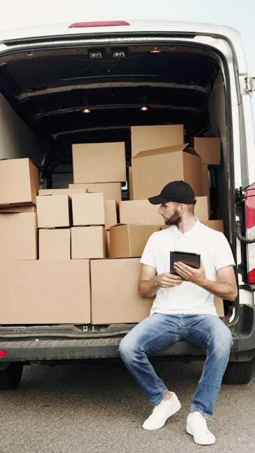 A man wearing a black cap, white T-shirt, and blue jeans is sitting on the pavement next to an open moving van during a home relocation in Kentish Town. The van's interior is filled with stacked cardboard boxes of various sizes, some sealed with packing tape and others partially open, indicating packing and moving preparation. The boxes are arranged on the floor of the van and against the side walls, with some overlapping. The man is holding a tablet device, possibly coordinating the loading process or documenting the move. The background shows a paved area outside the property, and the lighting suggests daylight. Visible equipment includes the moving van's interior structure, with metal rails and a dark ceiling. This scene reflects residential furniture transport and the packing and loading process associated with same-day removals services provided by Man with Van Kentish Town.