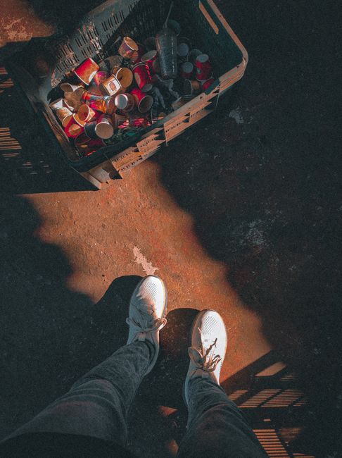 A person standing outdoors on a dirt surface, wearing white sneakers and dark jeans, with the shadow of a railing cast nearby. Next to their feet is a black plastic crate filled with used, empty aluminium cans, some with red and silver labels. The crate is positioned close to a doorway or loading area, suggesting a scene related to waste disposal or recycling as part of a home removal or moving process, which may be coordinated by Man with Van Kentish Town, a business specialising in removals and relocation services.