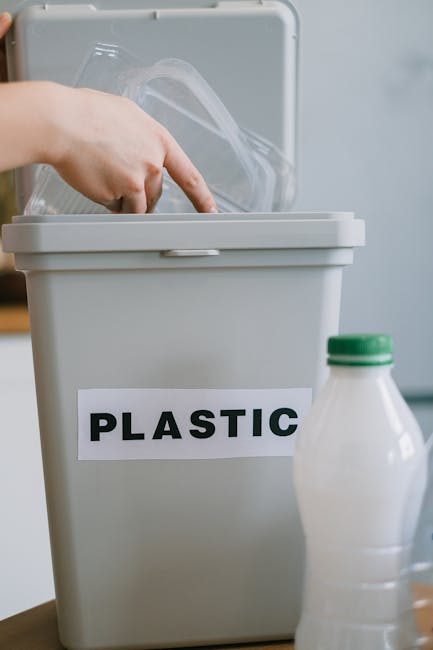 A grey plastic recycling bin with a white label reading 'PLASTIC' attached to its front, positioned on a wooden surface. The bin’s lid is open, revealing a crumpled transparent plastic bag inside. In front of the bin, there is a partially visible plastic bottle with a green cap. The setting appears to be indoors, with a neutral background. This image relates to waste disposal and recycling practices that are relevant during home removals or moves, supported by Man with Van Kentish Town’s house removal services, emphasizing proper packing and materials disposal during furniture transport and moving procedures.